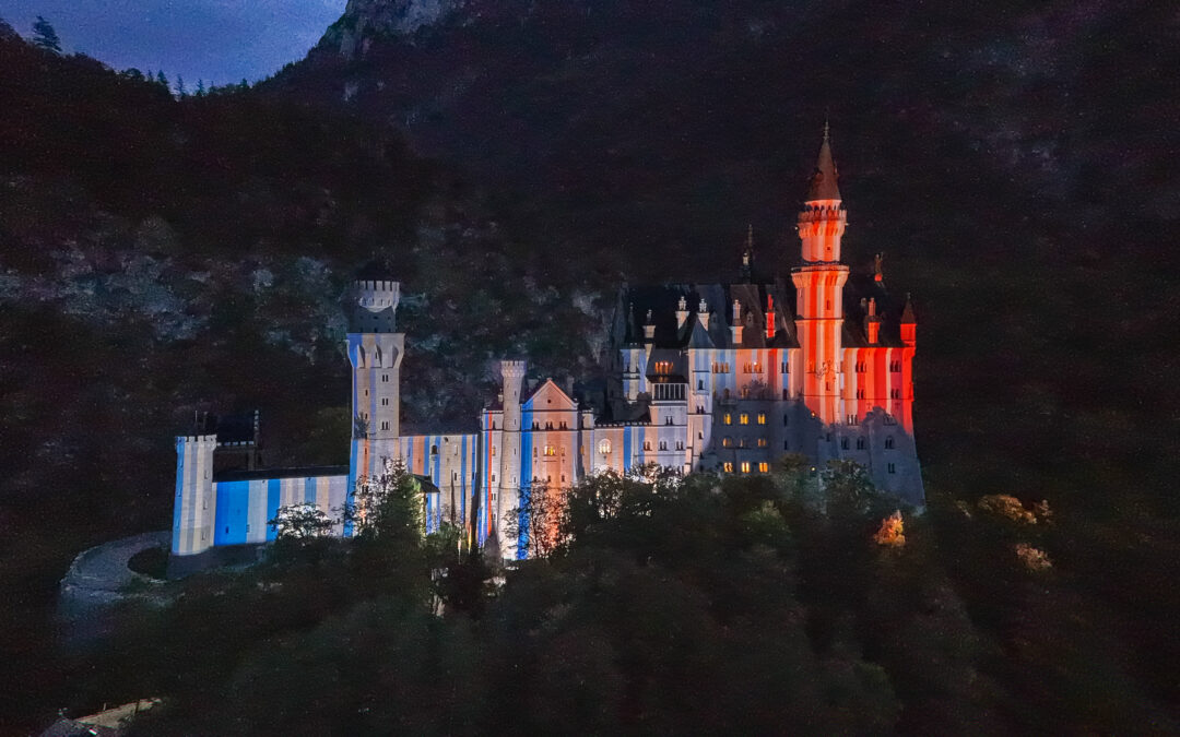 Das Schloss Neuschwanstein und auf dessen Fassade projeziert aufrechte Streifen, links eher blau, rechts eher rot, stark rot Nachthimmel
