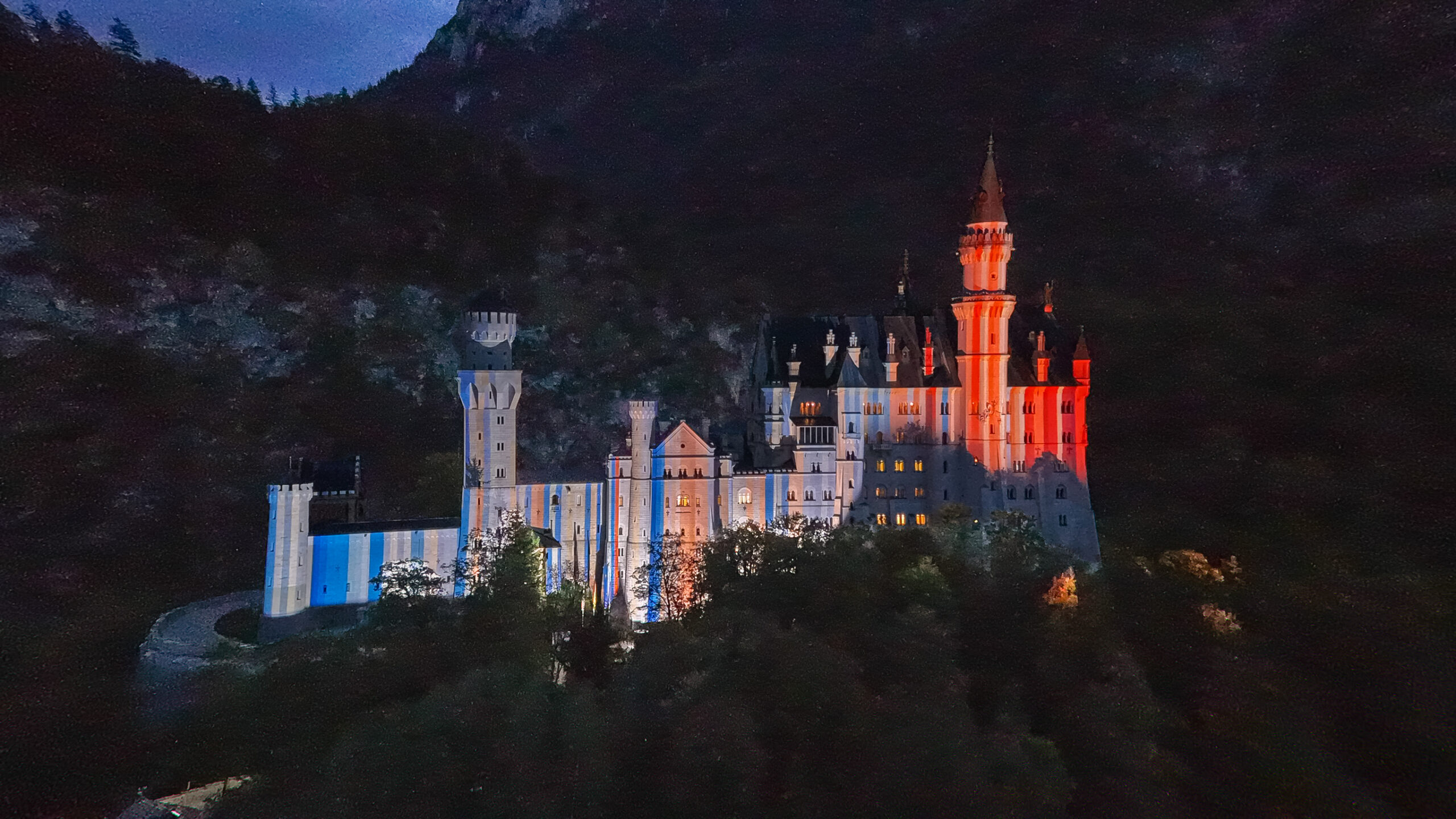 Das Schloss Neuschwanstein und auf dessen Fassade projeziert aufrechte Streifen, links eher blau, rechts eher rot, stark rot Nachthimmel